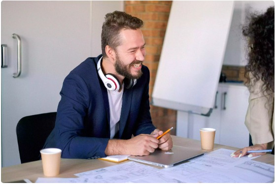 Smiling man wearing headphones at a desk with a laptop and coffee cups, engaged in discussion about online radio hosting services.