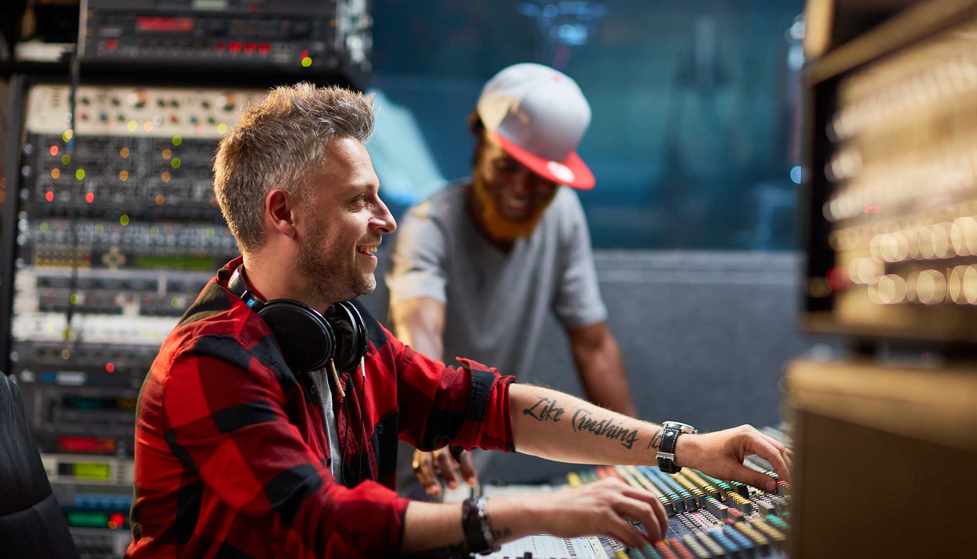 Two men in a radio studio, one adjusting audio-mixer controls while wearing headphones and a red plaid shirt, the other smiling in the background, emphasizing CastHost's secure radio-streaming services.